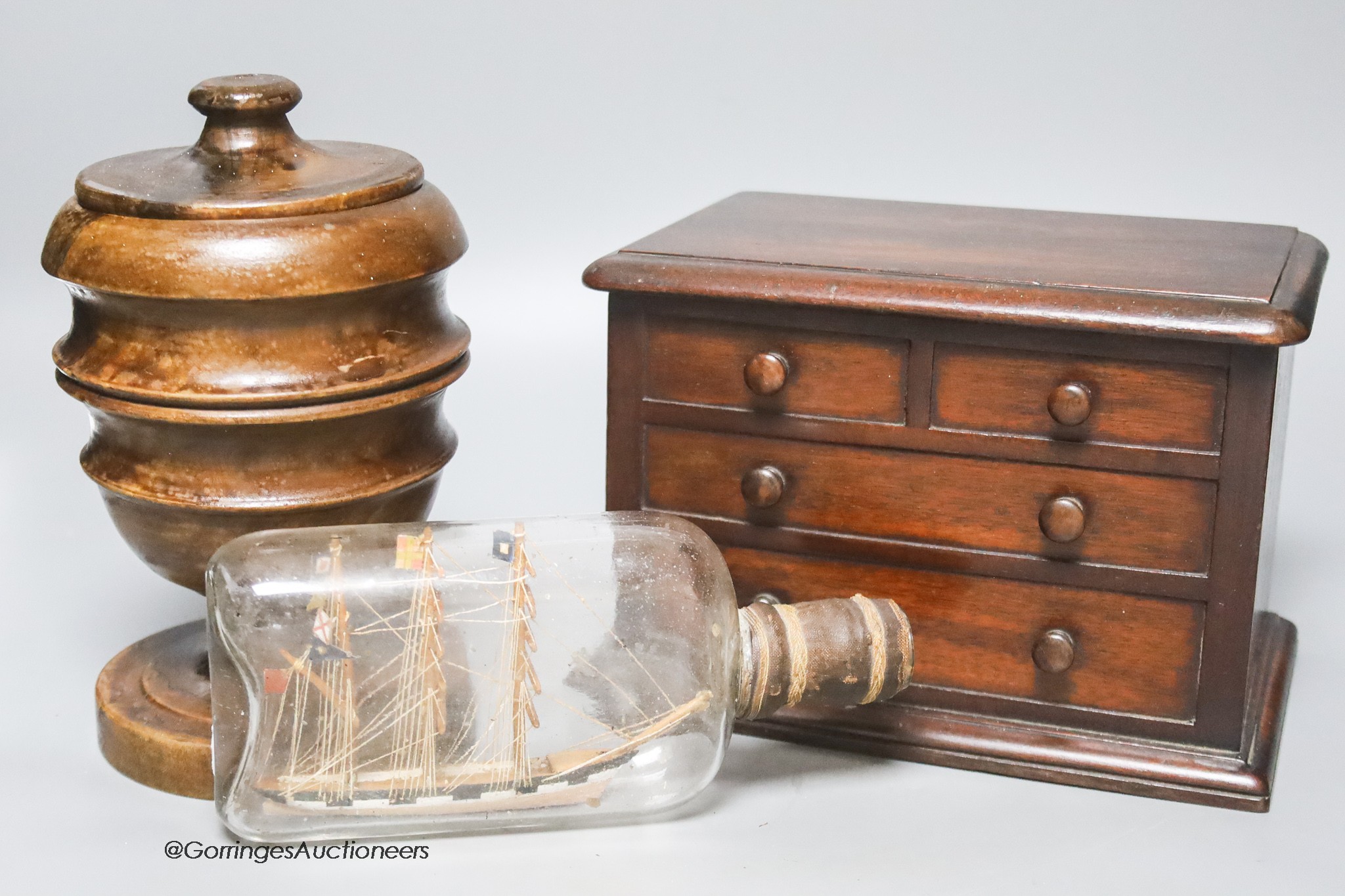 A Victorian mahogany 'chest of drawers' money bank, a turned wood bowl and cover and a small ship in a bottle, tallest 19cm